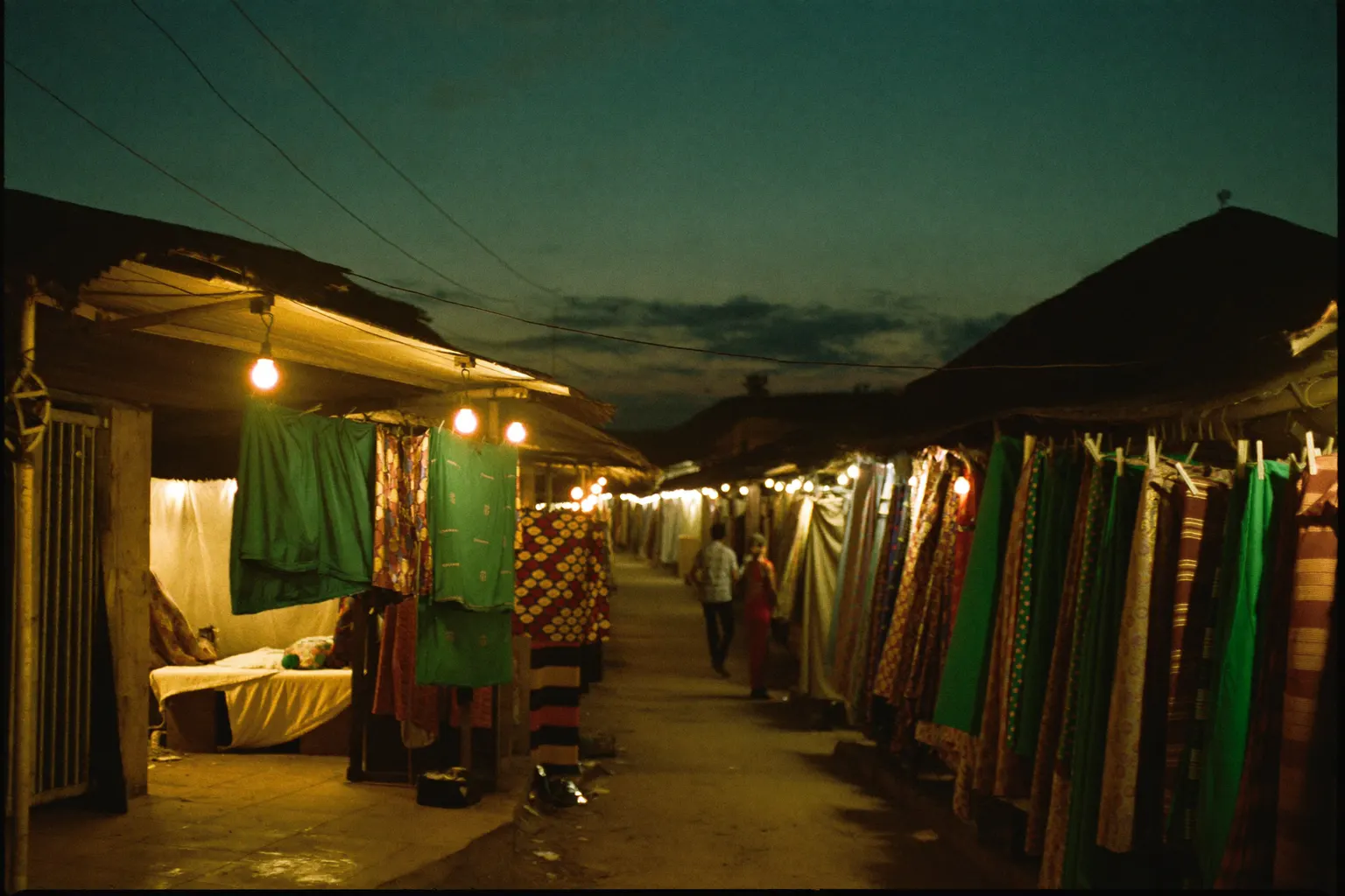 A fabric market street in Accra at dusk: stalls hung with kente and wax-print cloth, a tailor's open atelier spilling warm light onto the street under a deep blue-green sky