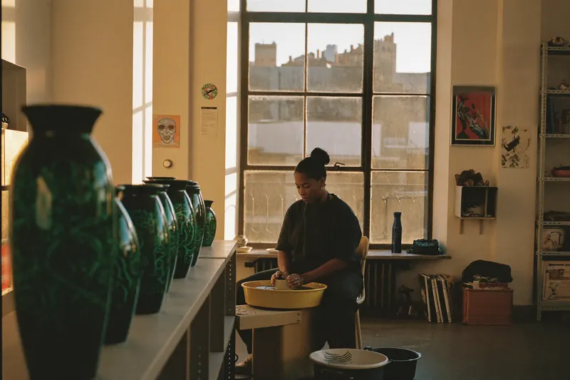 A ceramicist of the diaspora at her potter's wheel in a loft studio, shelves of emerald-glazed vessels behind her and low evening light through tall windows — an AI-generated plate