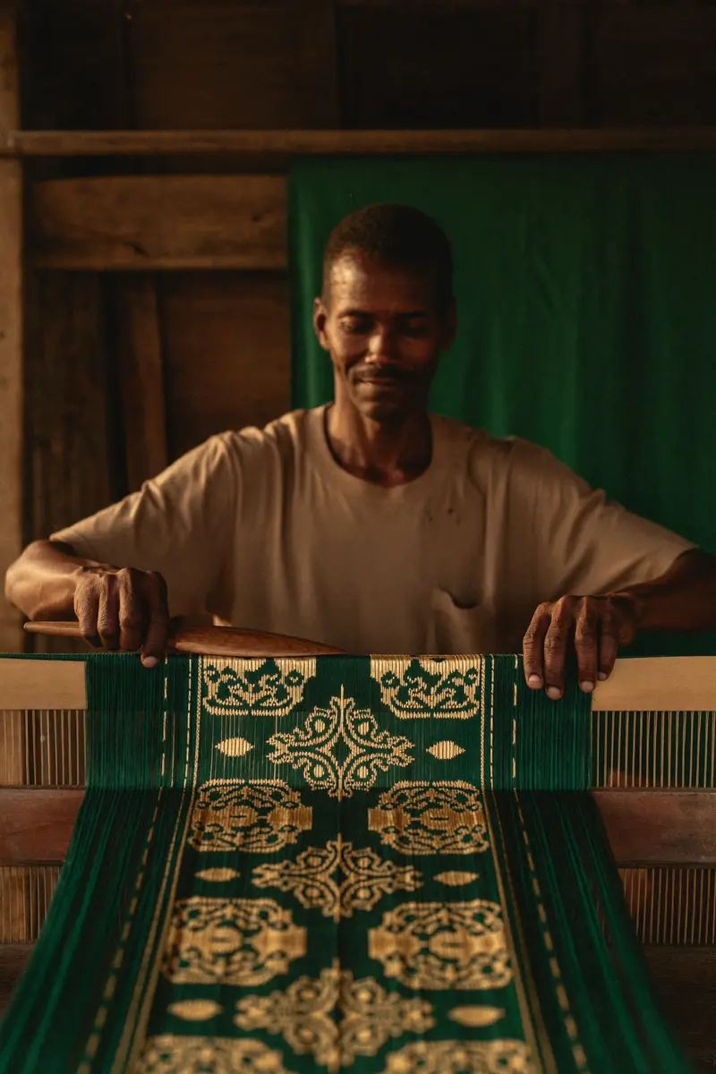 A kente weaver at his wooden strip loom, shuttle mid-throw through taut warp threads as an emerald-and-gold strip forms — an AI-generated portrait of the illustrative persona Yaw